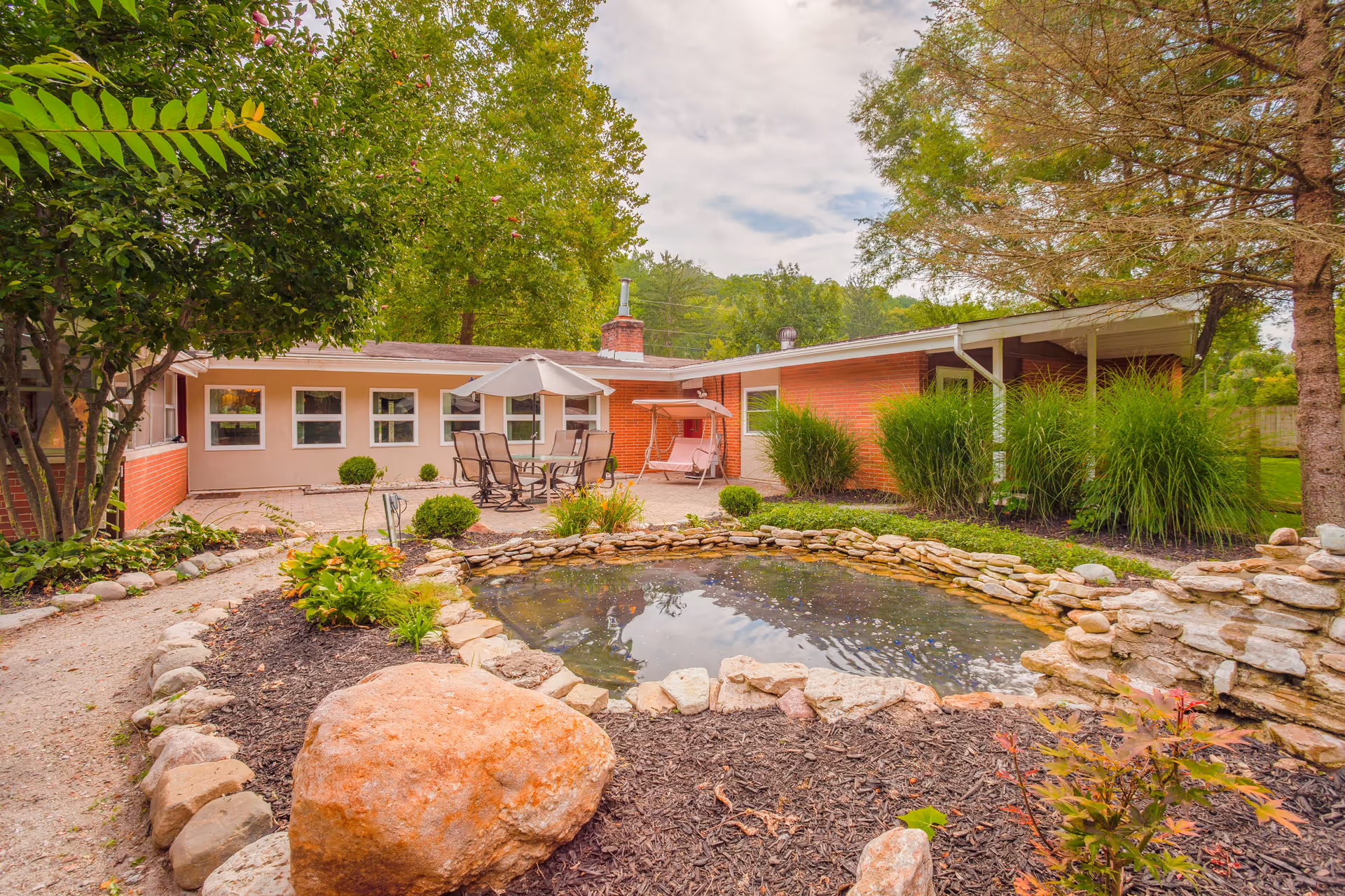 Outdoor garden area at Family Tree - Washington Township featuring a small pond surrounded by rocks and mulch, a patio with a table, chairs, and an umbrella, a pink swing, and lush greenery including trees and bushes.