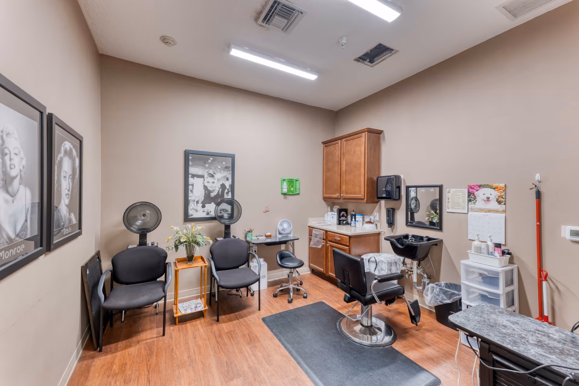 Interior of a small salon room with two black chairs under hair dryers, a salon styling chair in front of a black sink, wooden cabinets, a countertop with various supplies, and framed black and white portraits on the wall.