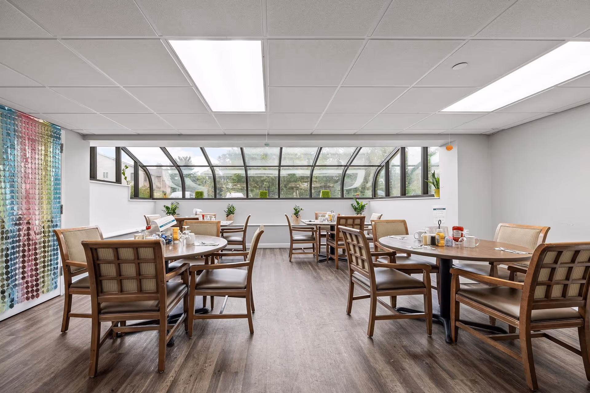 Bright dining room with round wooden tables and chairs set for meals beneath a curved skylight window.
