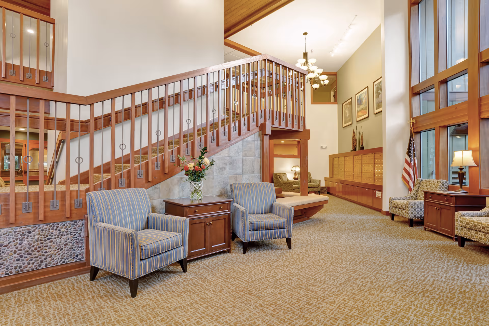 A spacious and well-lit interior common area featuring a wooden staircase with metal railings, two striped armchairs with a wooden side table and a vase of flowers between them, patterned carpet flooring, large windows with wooden frames, an American flag, and additional seating with patterned armchairs and a lamp on a wooden table.
