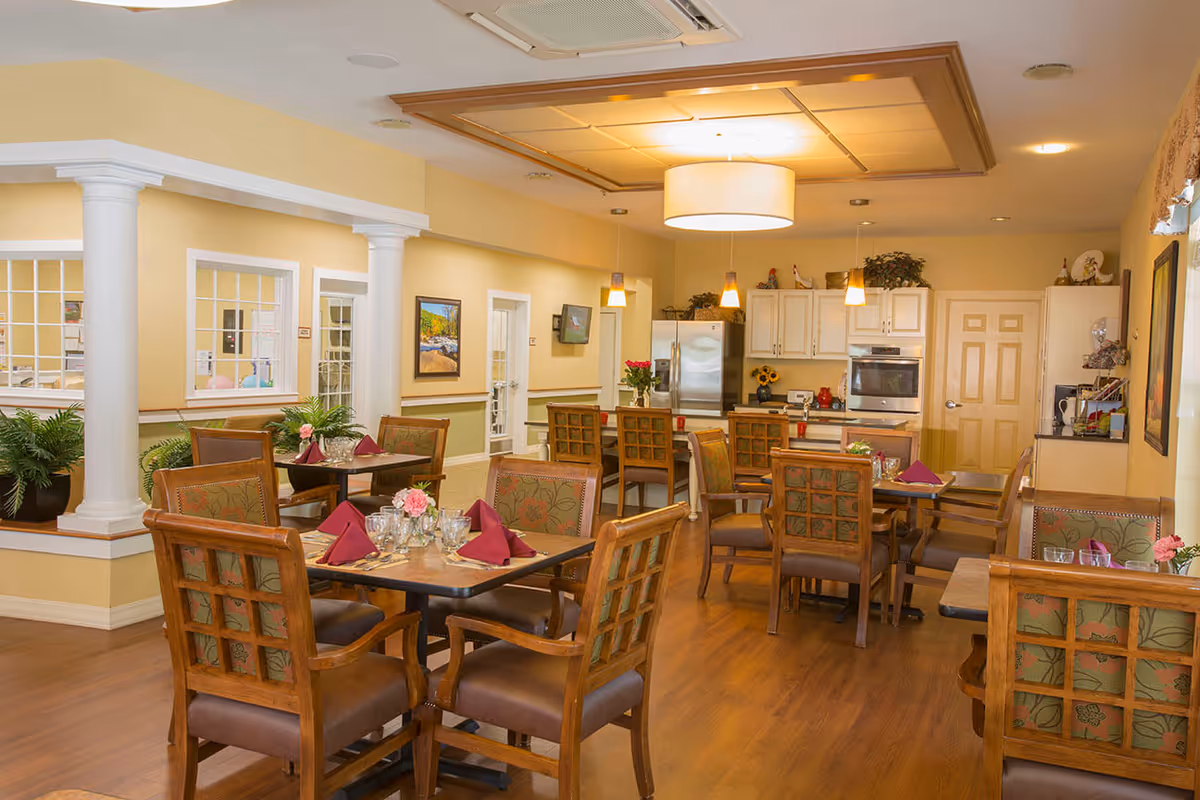 A cozy dining area in a senior living facility featuring wooden tables and chairs with floral upholstery. Tables are set with glasses, silverware, and burgundy napkins. The room has warm yellow walls, wooden flooring, white columns, and a kitchen area with white cabinets, a refrigerator, and an oven in the background. Soft lighting fixtures hang from the ceiling, creating a welcoming atmosphere.