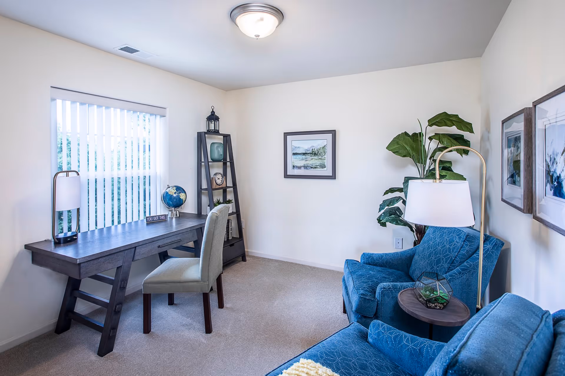 A bright and cozy living room with a large window covered by vertical blinds. There is a dark wooden desk with a lamp, a globe, and a small sign that says 'YOU GOT THIS'. A light gray upholstered chair is placed at the desk. In the corner, there is a tall black ladder shelf with decorative items including a clock and a lantern. Two blue upholstered armchairs are positioned near a small round side table with a lamp and a small plant. The walls are decorated with framed artwork and a large green plant adds a touch of nature to the room.