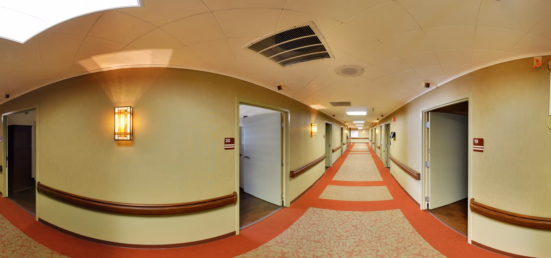 A long carpeted corridor inside a senior living facility with open room doors, wooden handrails, wall sconces, and recessed ceiling lights.