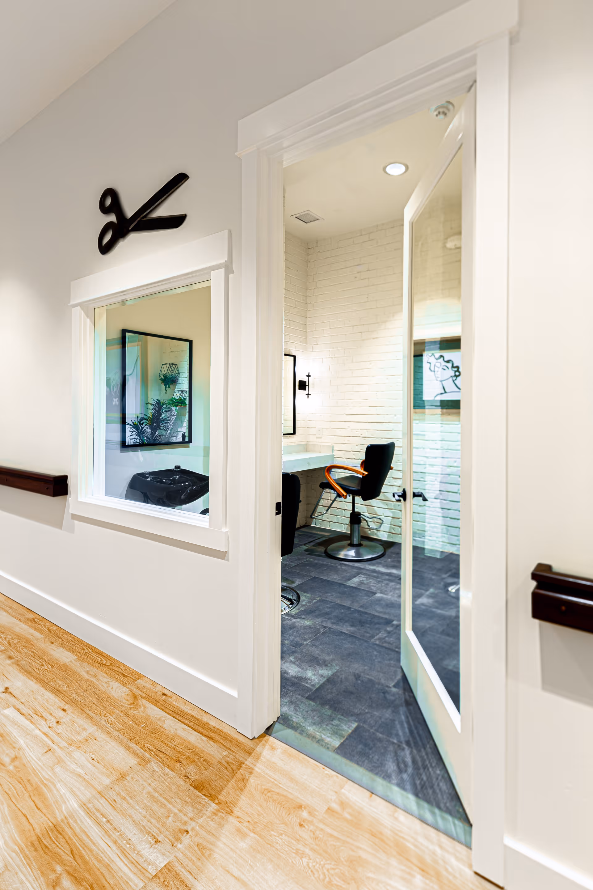 Interior view of a hair salon room in an assisted living facility. The room has a black salon chair, a white brick wall, a mirror, and a small counter. Outside the room, there is a hallway with wooden flooring and a wall decoration shaped like a pair of scissors above a window.