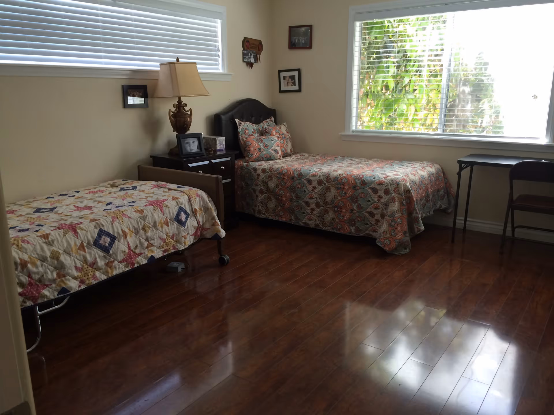 Simple bedroom with two twin beds, a nightstand and lamp, hardwood floors, and a large window with blinds.