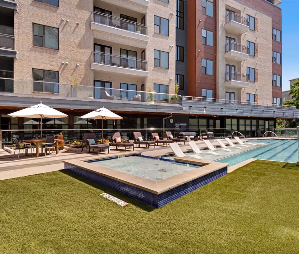Outdoor pool area at The Doliver of Tanglewood featuring a rectangular swimming pool with lounge chairs partially submerged in the water, a hot tub, several lounge chairs with cushions, tables with umbrellas, and a multi-story residential building in the background.