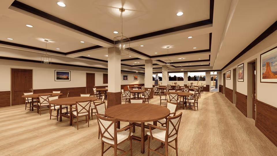 A spacious dining room with multiple round wooden tables and chairs arranged neatly. The room features a light wood floor, cream-colored walls with wooden wainscoting, framed landscape pictures on the walls, and a ceiling with recessed lighting and decorative beams. Large windows at the far end let in natural light.