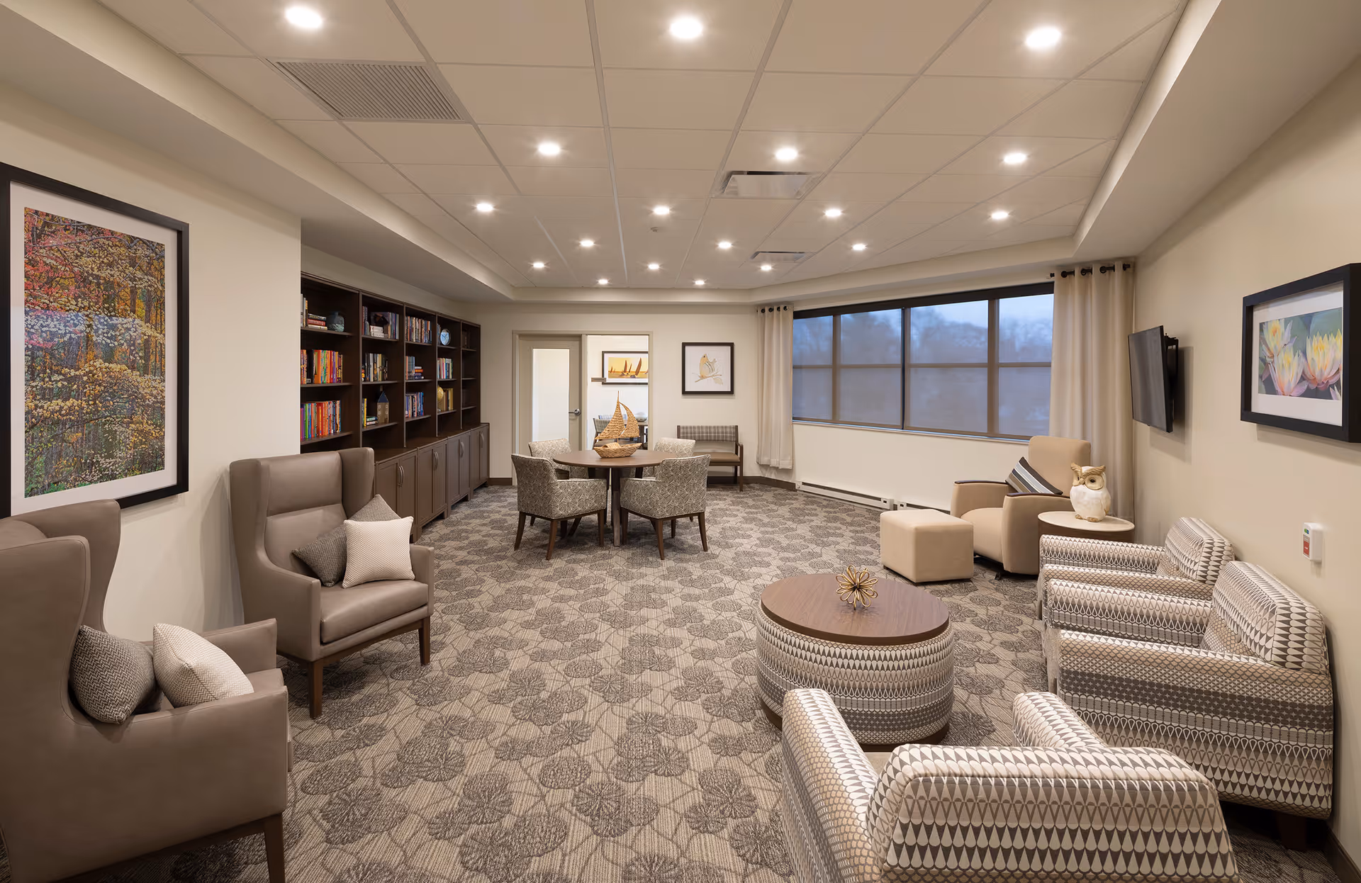 A well-lit communal lounge with upholstered chairs, round tables, bookshelves, a wall-mounted TV, and large windows.