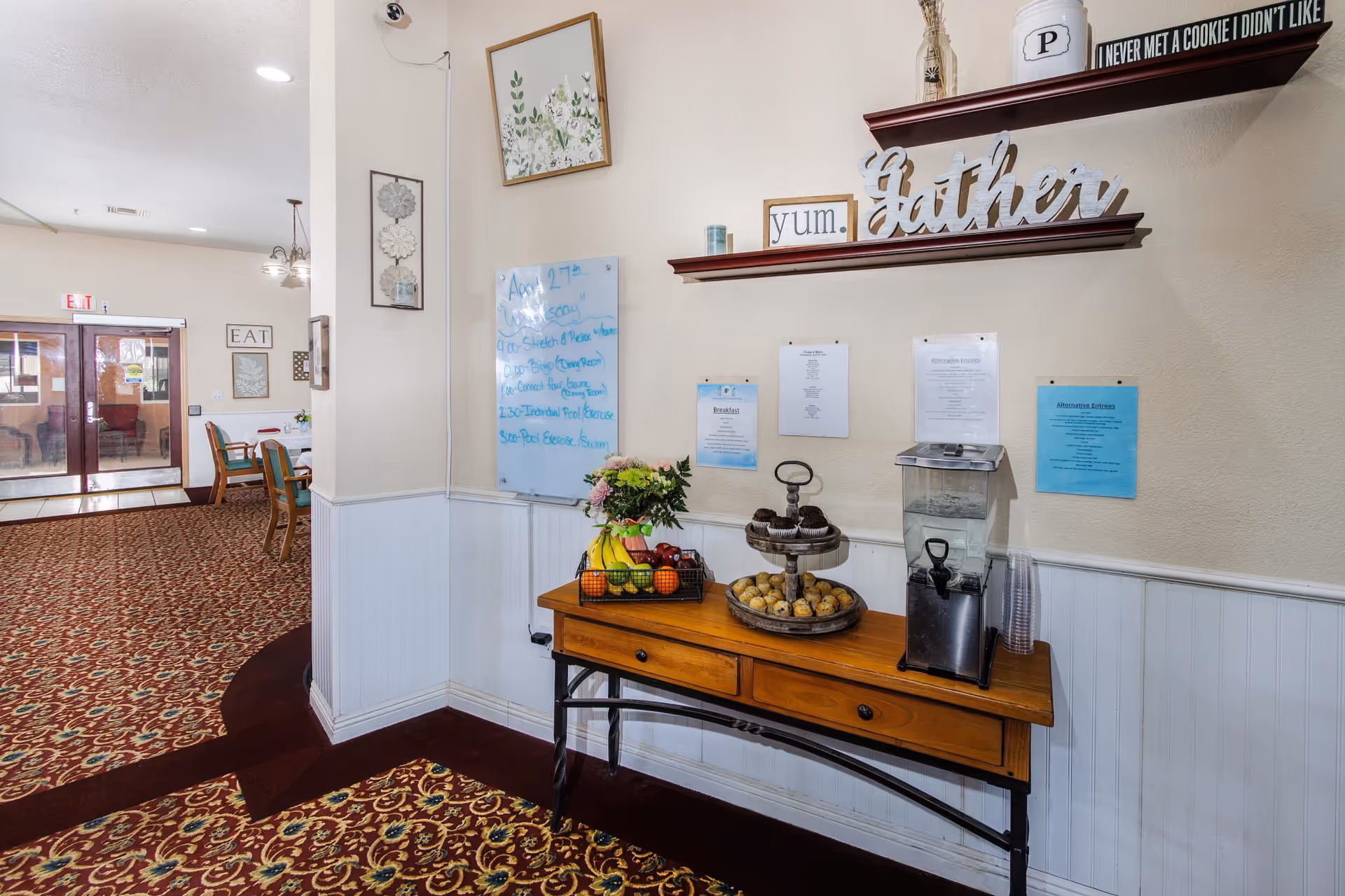 Interior view of a senior living facility hallway with a wooden table holding a tiered tray of muffins and bagels, a basket of fruit, a water dispenser with plastic cups, and a flower arrangement. Above the table are two wooden shelves with decorative items including a sign that says 'Gather' and a small sign that reads 'yum.'. On the wall behind the table are several posted notices and a whiteboard with handwritten notes. The hallway leads to a dining area with tables and chairs, visible in the background.