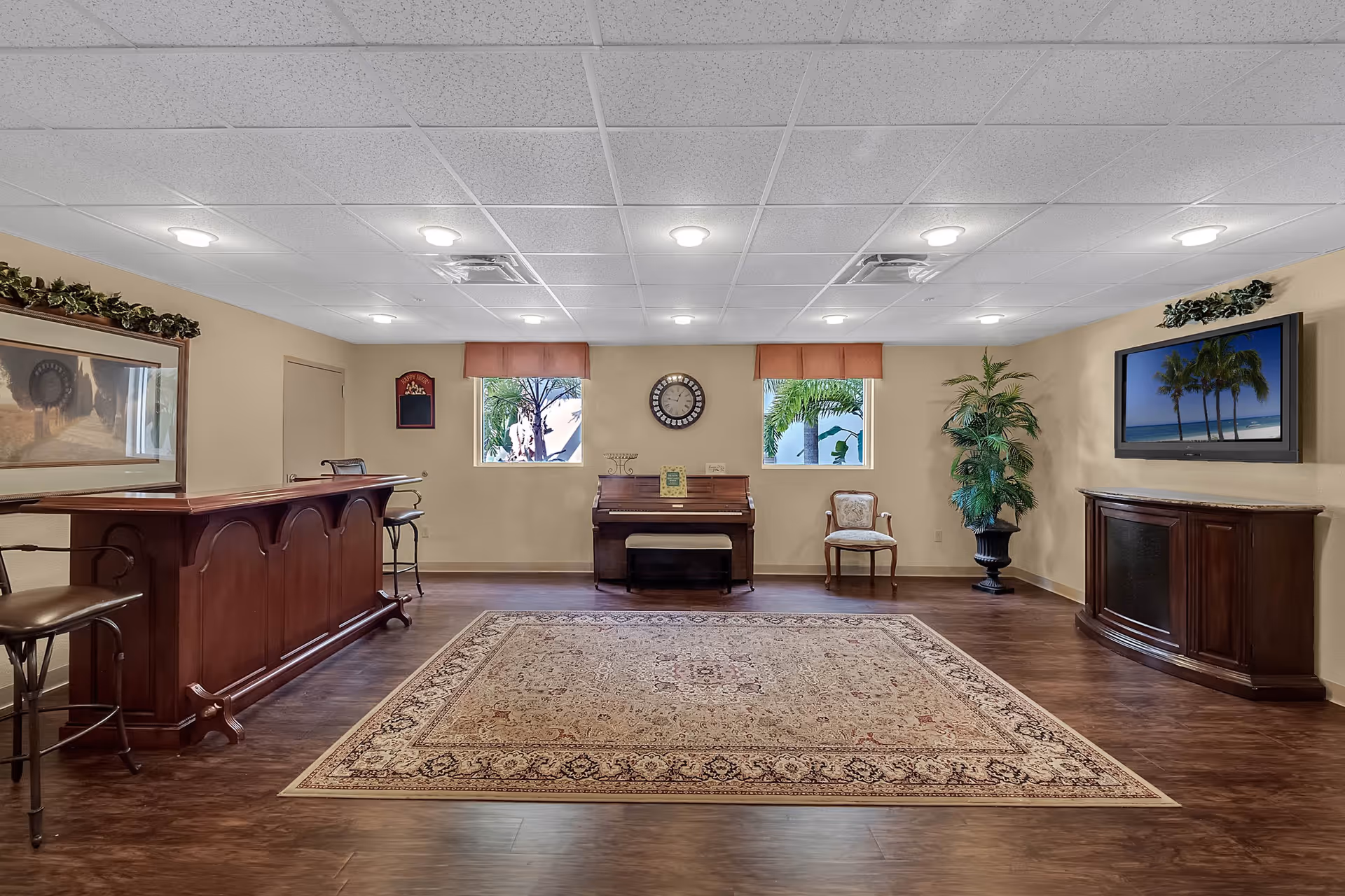 A spacious room with wooden flooring and a large patterned area rug in the center. The room features a wooden bar with stools on the left, a piano against the back wall between two windows with orange valances, a single upholstered chair, a large potted plant, and a flat-screen TV mounted on the right wall displaying an image of palm trees on a beach. The ceiling has recessed lighting and a drop ceiling with white tiles.