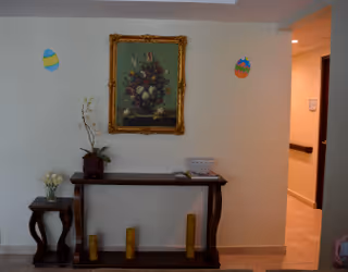 Interior hallway area with a wooden console table against a beige wall. On the table are a potted orchid, a basket, and some small items. Above the table hangs a framed painting of a floral arrangement. Two colorful egg-shaped decorations are attached to the wall on either side of the painting. The floor is tiled and a hallway extends to the right.