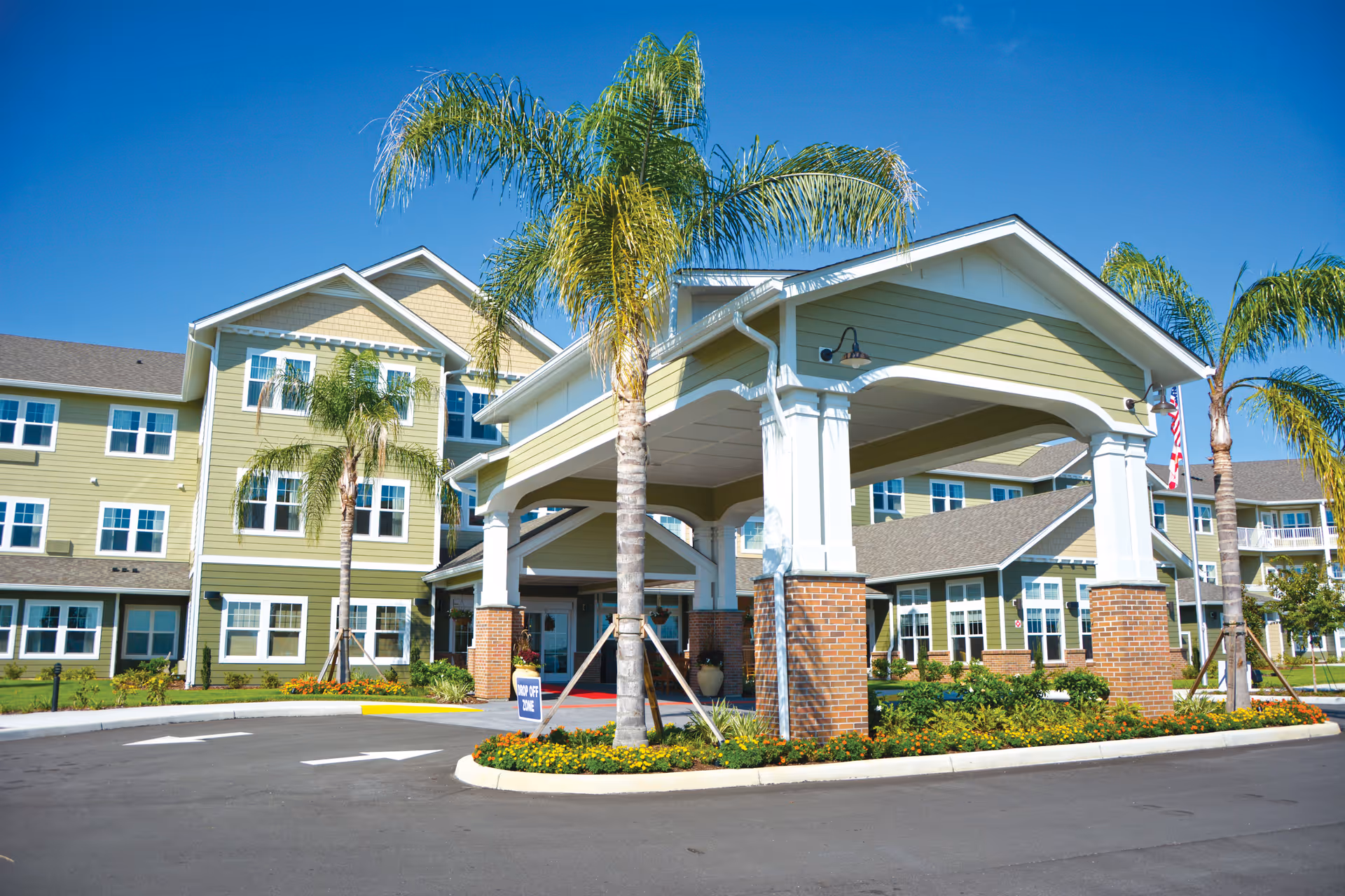 Front exterior of a multi-story senior living building with a covered porte-cochère, palm trees, and a landscaped driveway.