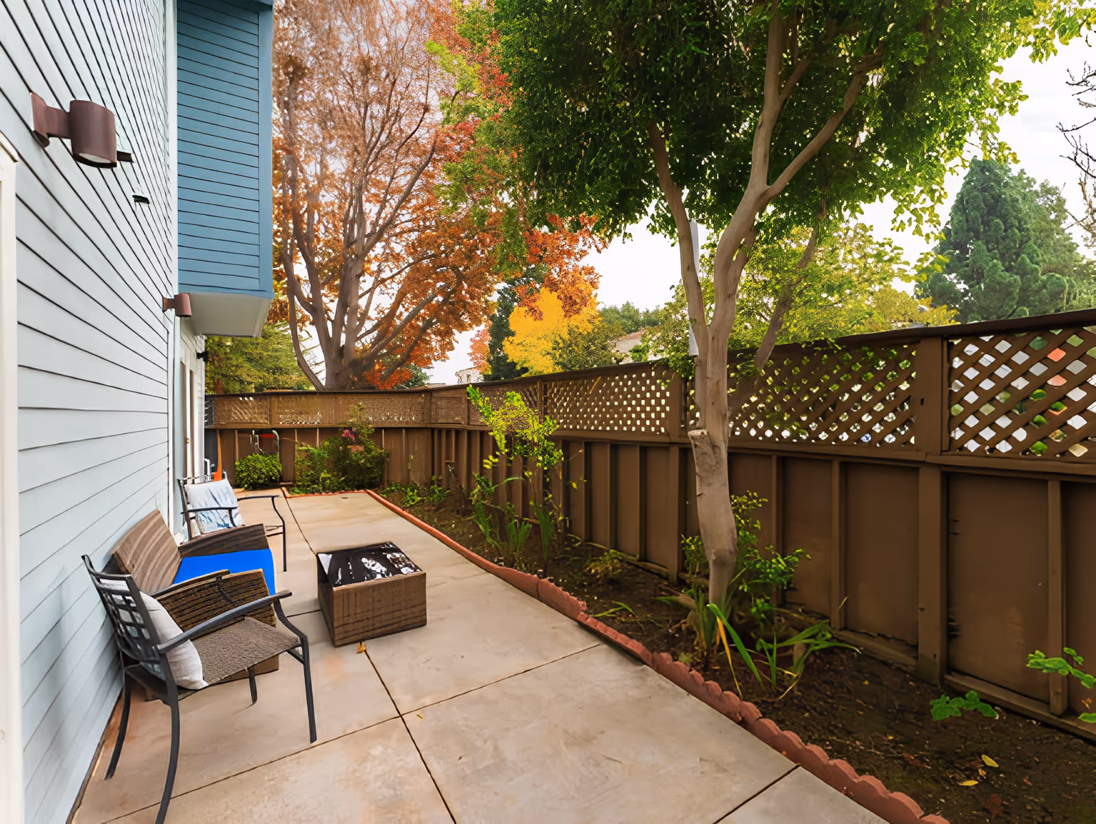 Outdoor patio area with a concrete floor, several chairs with cushions, a small table, and a brown wooden fence with lattice panels. There are trees with green and autumn-colored leaves surrounding the patio.
