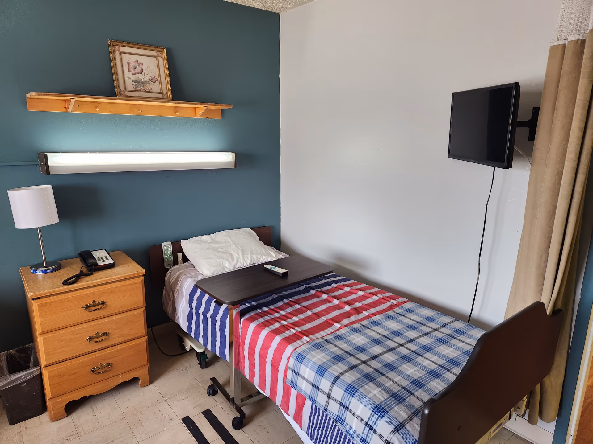 A small bedroom in a senior living facility with a single bed covered in a red, white, and blue plaid blanket. There is a wooden bedside table with a lamp, a telephone, and a remote control on the bed. A flat-screen TV is mounted on the wall opposite the bed. The walls are painted teal and white, with a wooden shelf holding a framed picture above the bed.