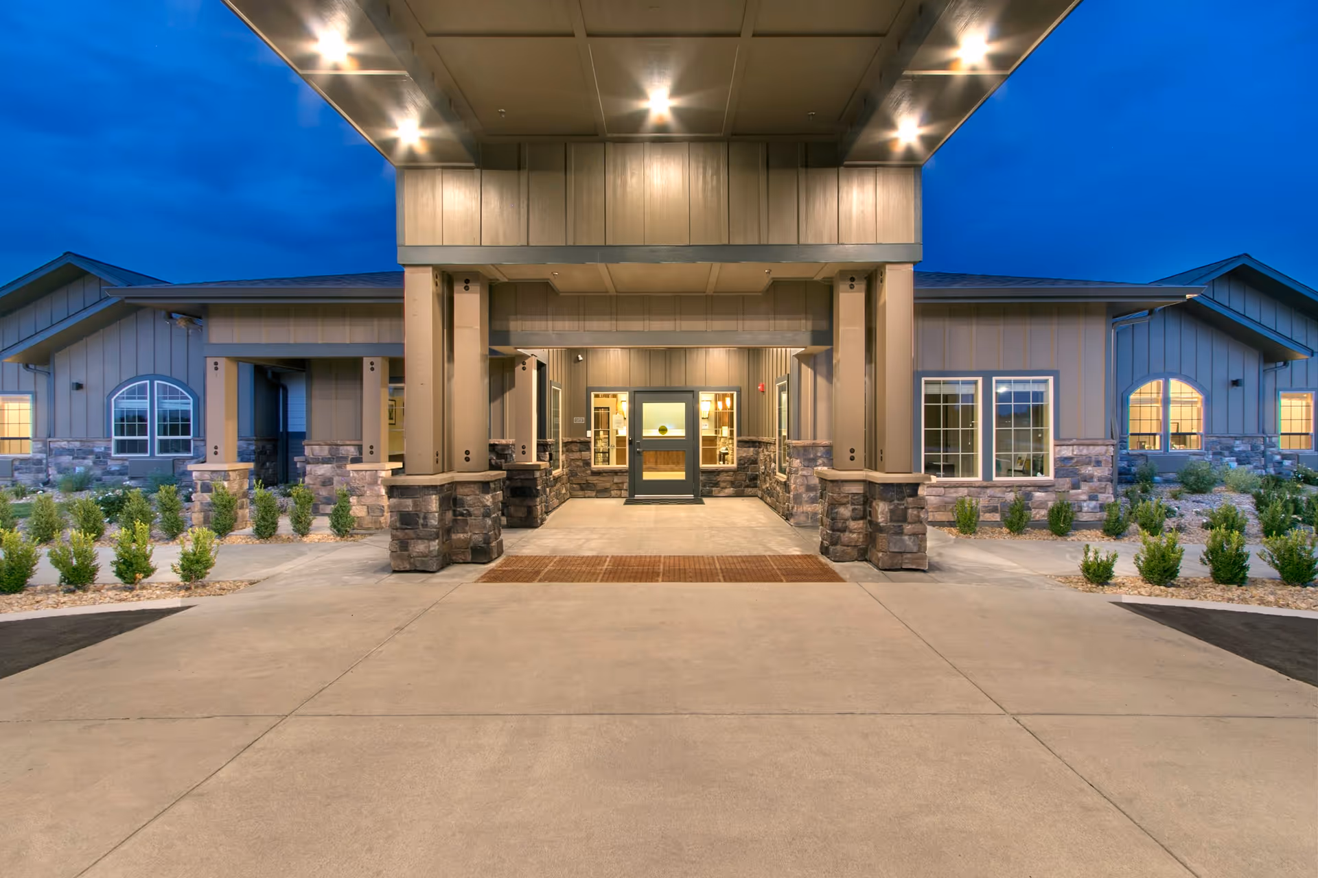 Front entrance of a senior living facility with a covered porte-cochere, stone columns, and lit windows at dusk.