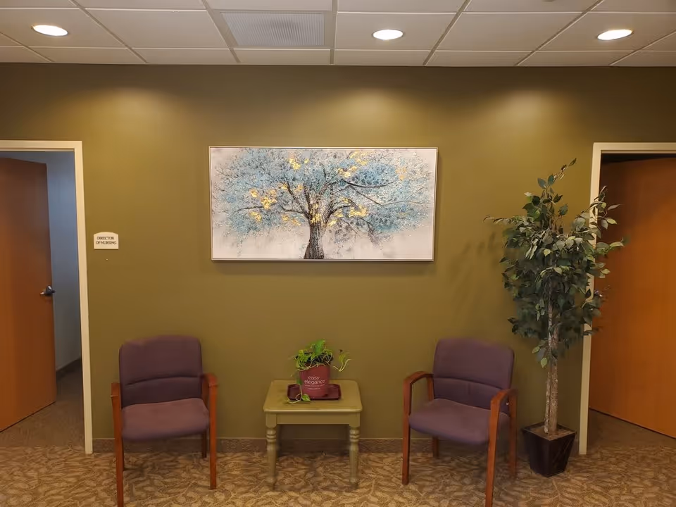 A waiting area with two purple cushioned chairs with wooden arms placed on either side of a small green table holding a potted plant. Behind the chairs is an olive green wall with a painting of a tree with blue and gold leaves. There is a tall potted plant to the right and two open wooden doors on either side of the wall. A small sign next to the left door reads 'Director of Nursing'.