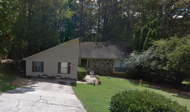 A single-story house with a slanted roof, beige siding, and stone facade on one side. The house is surrounded by green grass, bushes, and tall trees in the background. There is a concrete driveway leading up to the house.