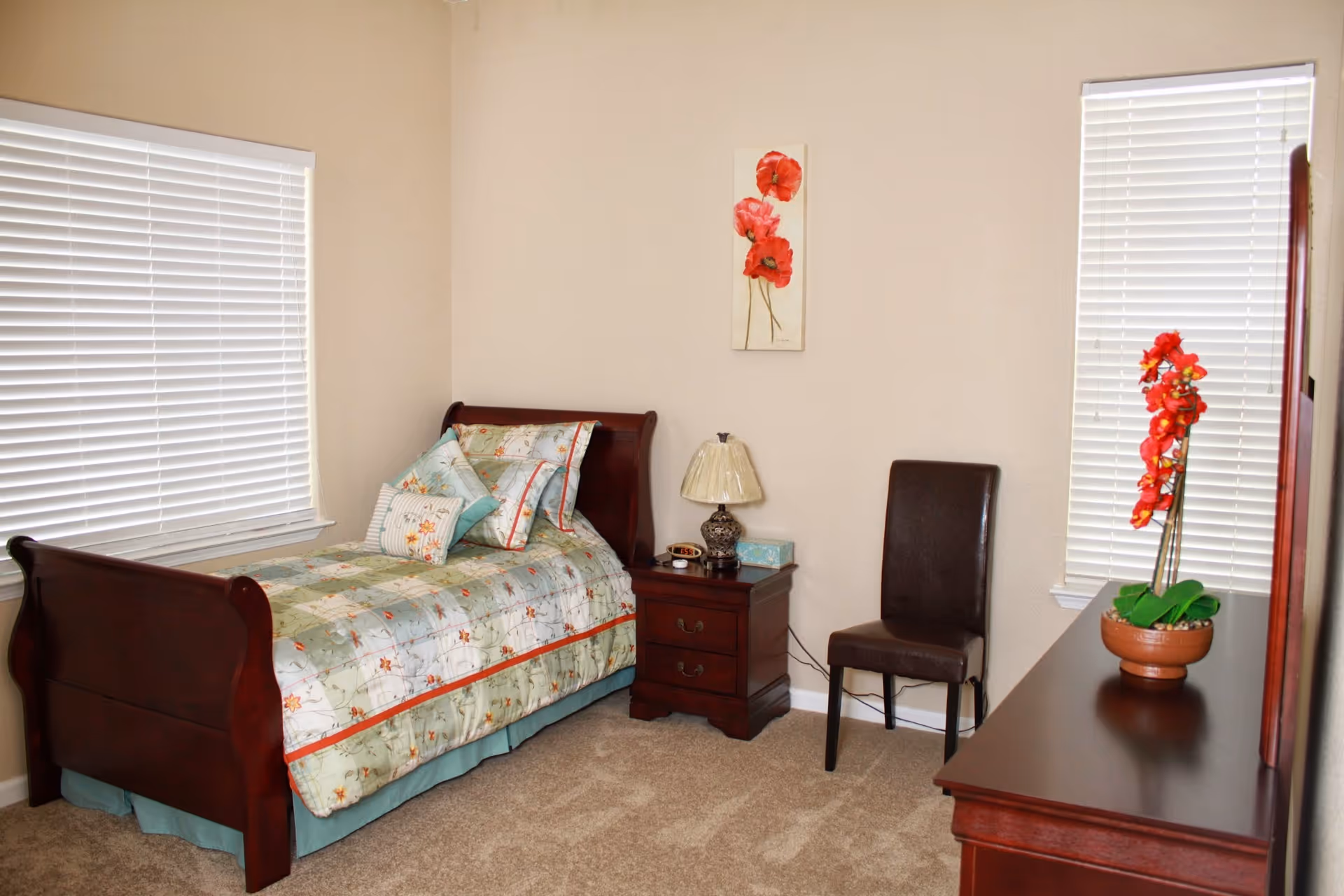 A neatly arranged bedroom with a single wooden bed featuring floral bedding and multiple pillows. Next to the bed is a wooden nightstand with a decorative lamp, alarm clock, and tissue box. A brown chair is placed beside the nightstand. The room has beige walls with a floral painting hanging above the nightstand, two windows with white blinds, and a wooden dresser with a potted red orchid on top.