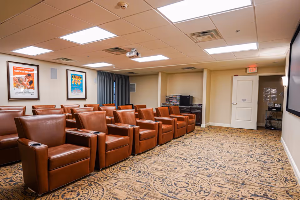 A small theater room with two rows of brown leather recliner chairs facing a large screen on the right wall. The room has patterned carpet, beige walls, a white ceiling with recessed lighting, and two framed movie posters on the left wall. A projector is mounted on the ceiling, and there is a cabinet with DVDs and electronic equipment at the back of the room.