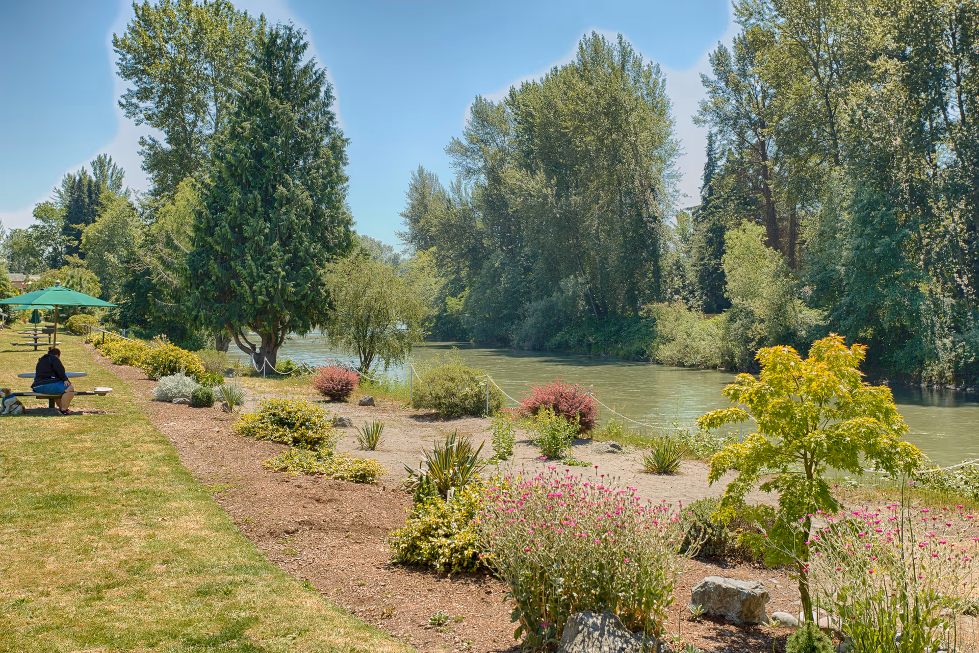 A peaceful outdoor area with a river running alongside a landscaped garden featuring various shrubs, small trees, and flowers. There are benches with green umbrellas and a person sitting on one of the benches. Tall trees line the opposite side of the river under a clear blue sky.