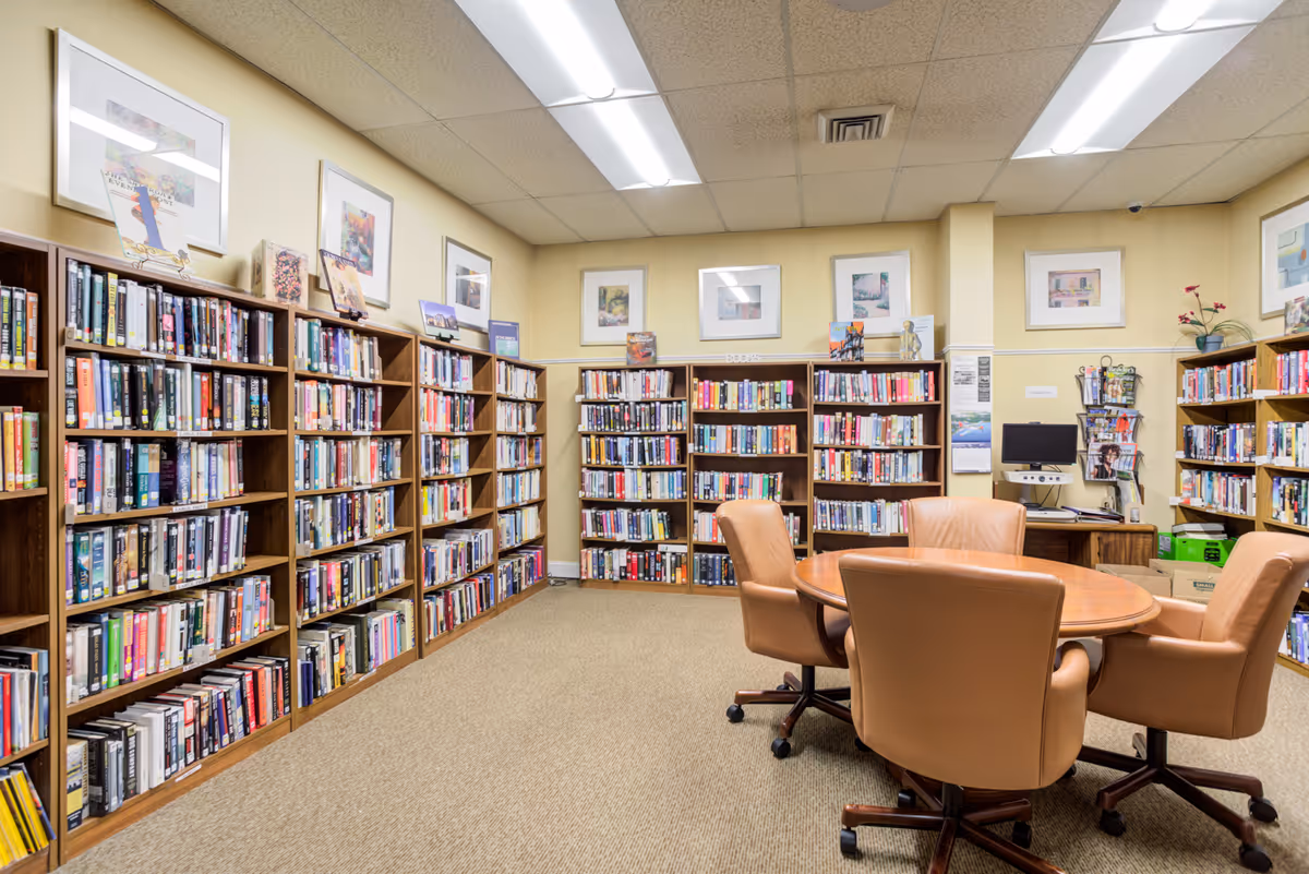 A well-lit library room with multiple wooden bookshelves filled with books along the walls. In the center, there is a round wooden table surrounded by four tan leather swivel chairs. The walls are painted light yellow and decorated with framed artwork. A computer workstation and magazine rack are visible in the back corner.