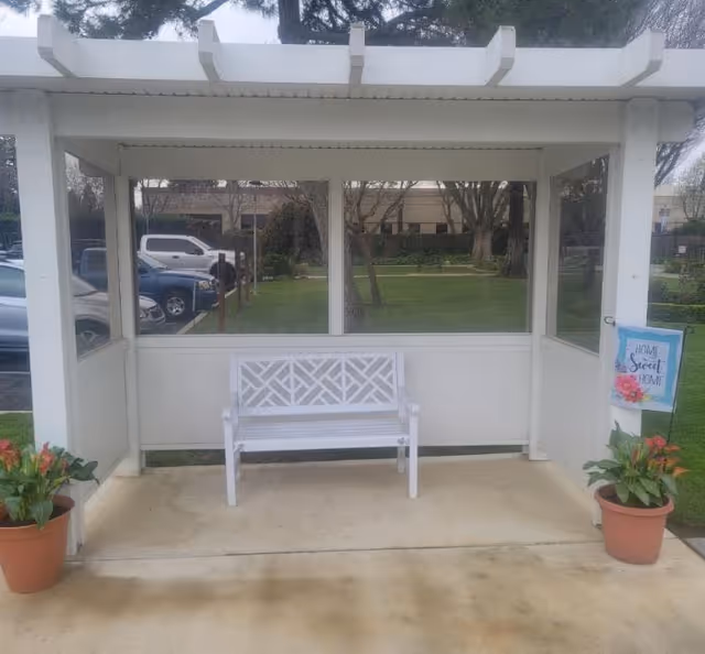 A white wooden bench under a white pergola structure with glass panels on three sides. Two potted plants with red flowers are placed on either side of the bench. A small decorative sign on the right side reads 'Home Sweet Home'. In the background, there is a green lawn with trees and parked cars visible.