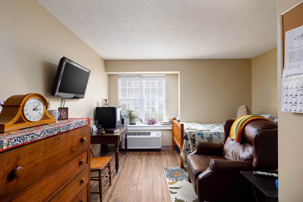 Small studio-style senior room with a bed, leather armchair, wooden dresser, wall-mounted TV and a window seat.