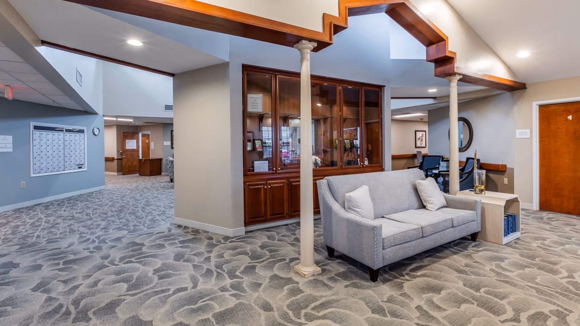Interior view of a senior living facility lobby area with patterned carpet, a gray sofa with two white pillows, a small side table with books and a decorative item, wooden columns, and a wooden display cabinet with glass doors. The area is well-lit with recessed ceiling lights and has light-colored walls and doors.