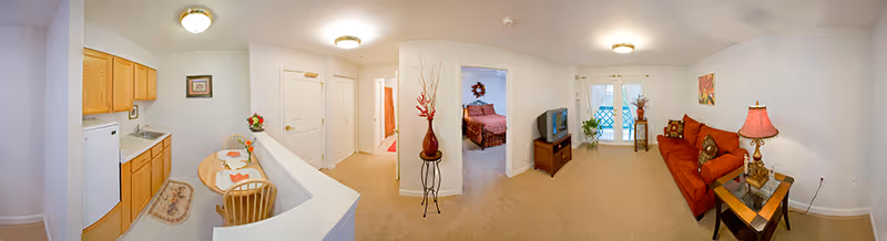 Panoramic interior view of a senior living apartment showing a small kitchen and dining area on the left, a hallway and bedroom in the center, and a living room with a sofa and TV on the right.