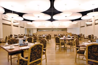 A spacious dining room with multiple tables and chairs arranged neatly. Each table is set with cups, saucers, and napkins. The room features large, round ceiling lights and light-colored wooden flooring.