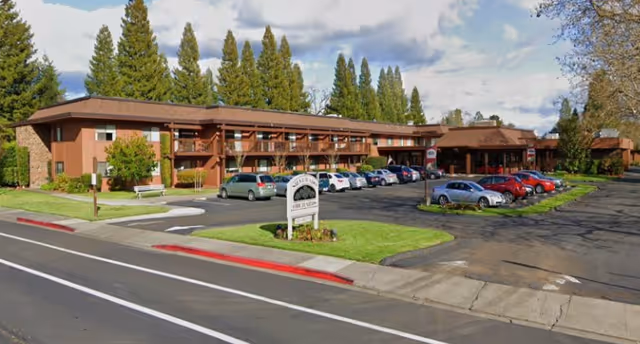 Exterior view of Silverado Orchards Retirement Community building with a parking lot in front, several parked cars, green lawns, trees in the background, and a sign displaying the facility name near the street.