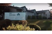 Exterior view of a single-story building with a sign in front that reads 'Foss' along with services offered such as Adult Day Care, Assisted Living, Memory Support, and more. The building is surrounded by grass, bushes, and trees.