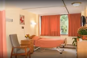 A simple, clean bedroom in a care facility with a single bed covered in a peach-colored blanket, a wooden nightstand with a flower vase, a gray chair, a dresser with a plant and a TV mounted on the wall, and a window with orange curtains letting in natural light.