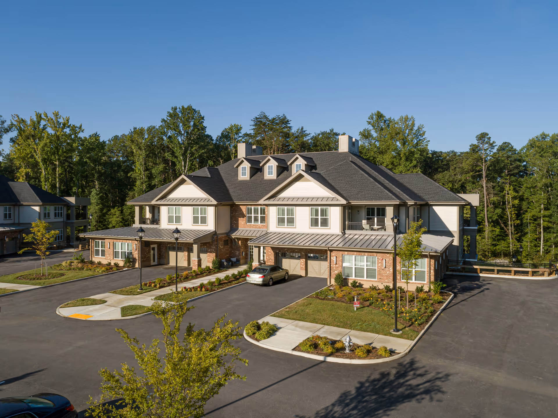 Exterior view of a two-story residential building at Lakewood Retirement Community surrounded by trees and landscaping, with a parking area and a few cars parked nearby under a clear blue sky.
