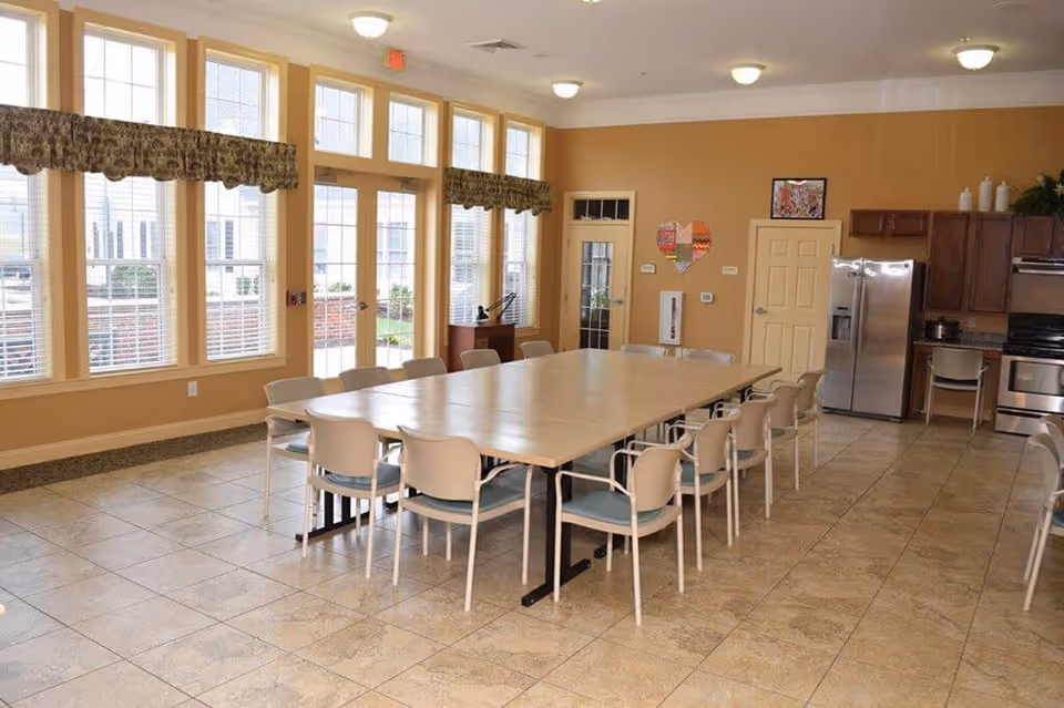 A spacious dining room with a large rectangular table surrounded by multiple chairs. The room has large windows with patterned valances allowing natural light to fill the space. In the background, there is a kitchen area with stainless steel appliances including a refrigerator and stove, wooden cabinets, and a tiled floor.