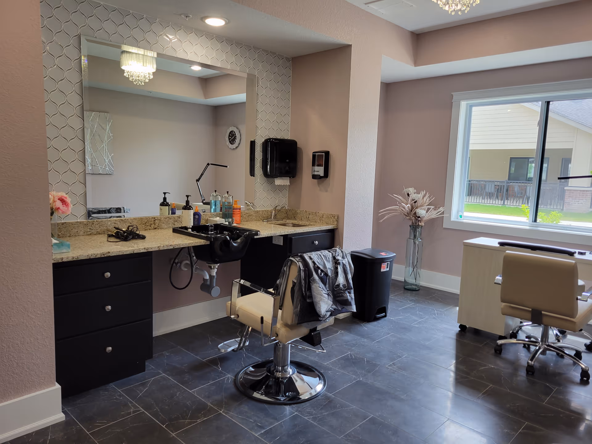 Interior view of a salon area in an assisted living facility featuring a styling chair in front of a large mirror with a granite countertop. Various hair care products and a hair washing sink are visible on the counter. There is a window letting in natural light, a trash bin, and a small desk with a chair near the window. The floor is tiled and the walls are painted light pink.