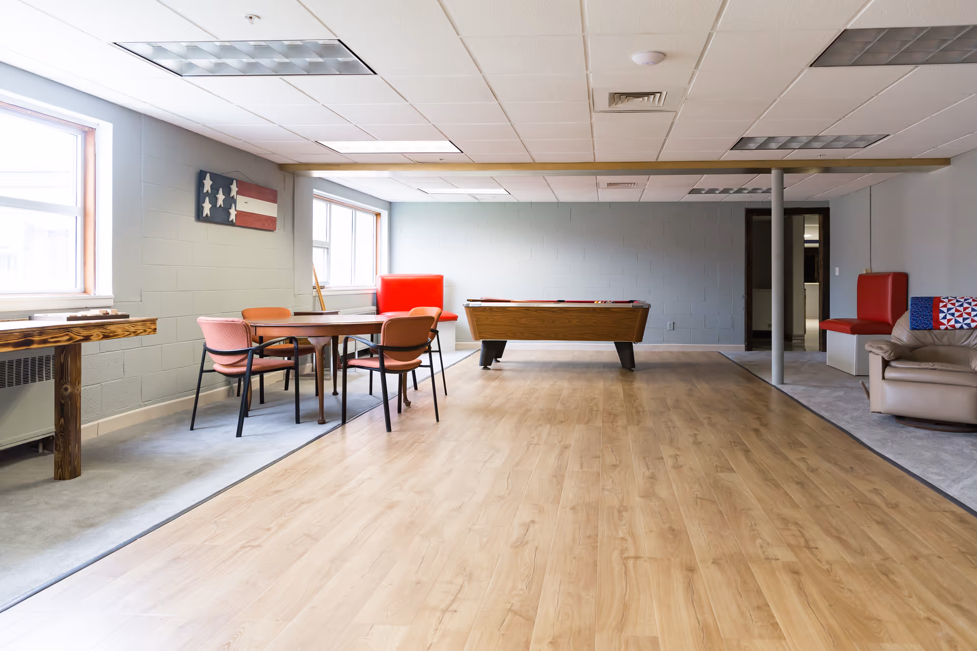 A spacious common room with light wood flooring and gray walls. The room features a pool table in the center, a wooden table with four chairs on the left, a red cushioned bench against the far wall, and a beige armchair with a colorful pillow on the right. Two large windows let in natural light, and there is a decorative wooden American flag on the wall.