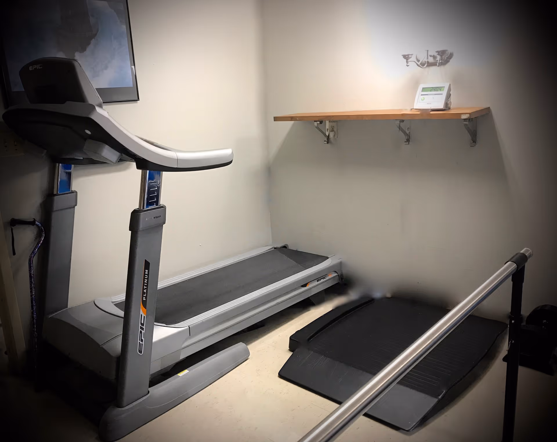Small exercise room with a treadmill, a black exercise mat, a wooden shelf mounted on the wall holding a small electronic device, and a metal handrail in the foreground.