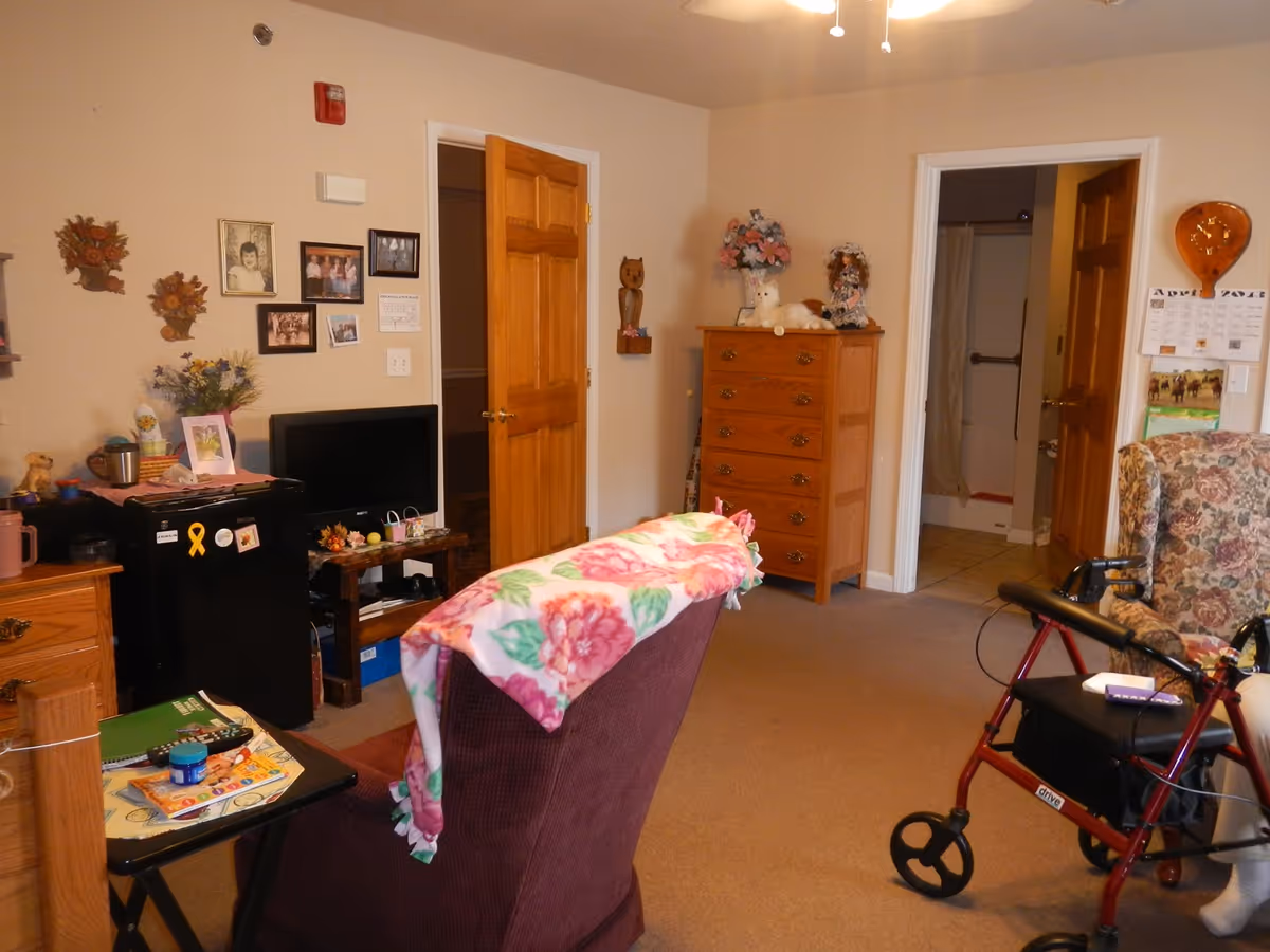 A cozy senior living room with an upholstered recliner draped in a floral blanket, a TV on a small stand, a wooden dresser, and a red rollator near an open doorway to a bathroom.