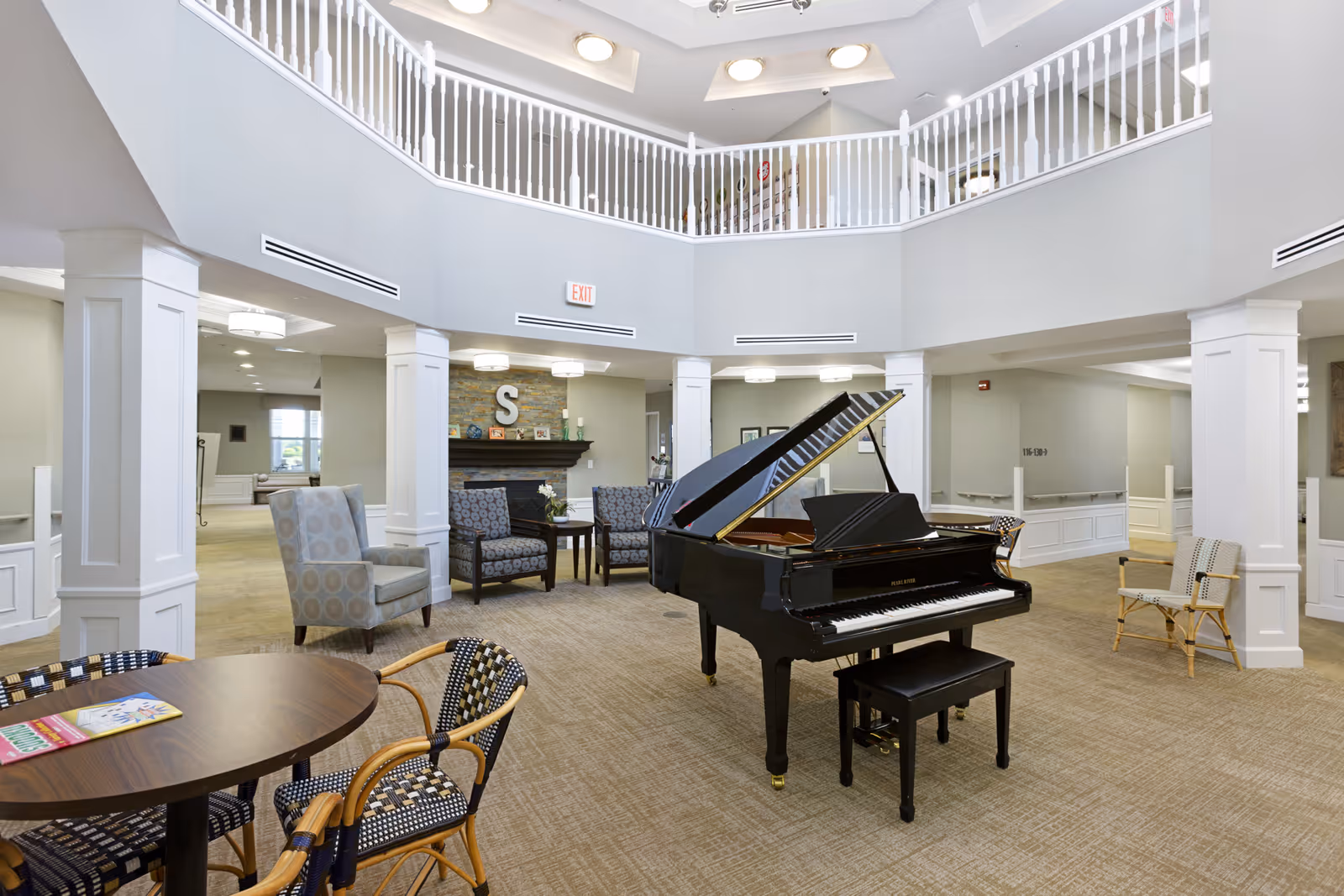 Spacious senior living facility common area with a black grand piano in the center, surrounded by various seating options including armchairs and wicker chairs. The room features a high ceiling with a balcony railing above, neutral-colored walls, carpeted floor, and a fireplace with decorative items on the mantel.