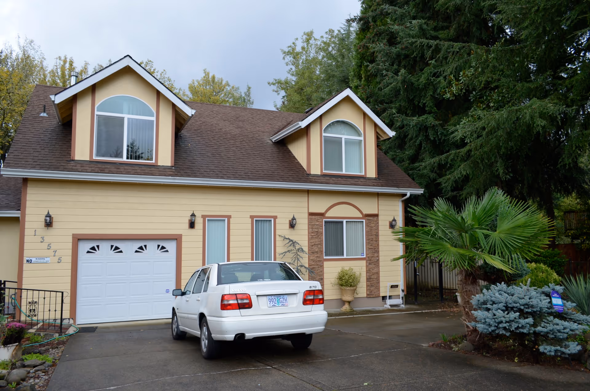 Two-story yellow house with a garage and a white car parked in the driveway, surrounded by trees and landscaping.
