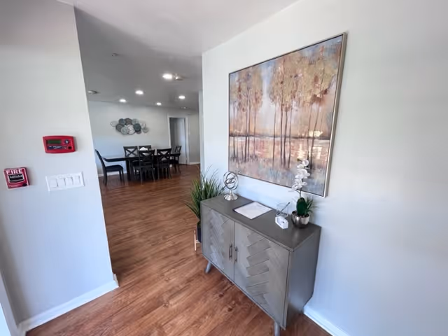 Interior view of a senior living facility hallway with wooden flooring and white walls. A decorative cabinet with plants and ornaments is placed against the right wall beneath a large framed painting of trees. In the background, there is a dining area with a dark wooden table and chairs. Ceiling lights illuminate the space.