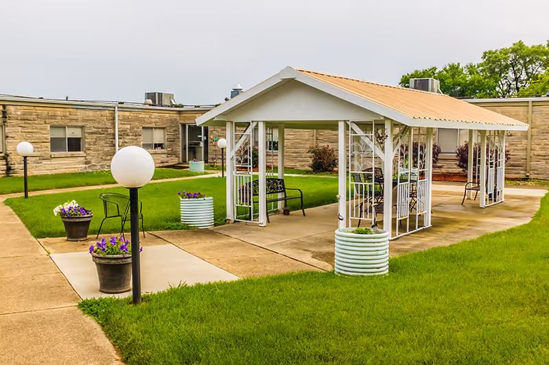 Outdoor courtyard area at a senior living facility with a covered pavilion featuring metal benches and chairs, surrounded by green grass, flower pots, and globe-shaped lamp posts. The building with beige brick walls and windows is visible in the background.