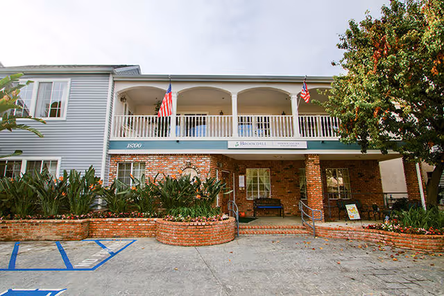 Front entrance of a two-story senior living building with a brick facade, covered balcony with American flags, planter beds, and parking spaces.