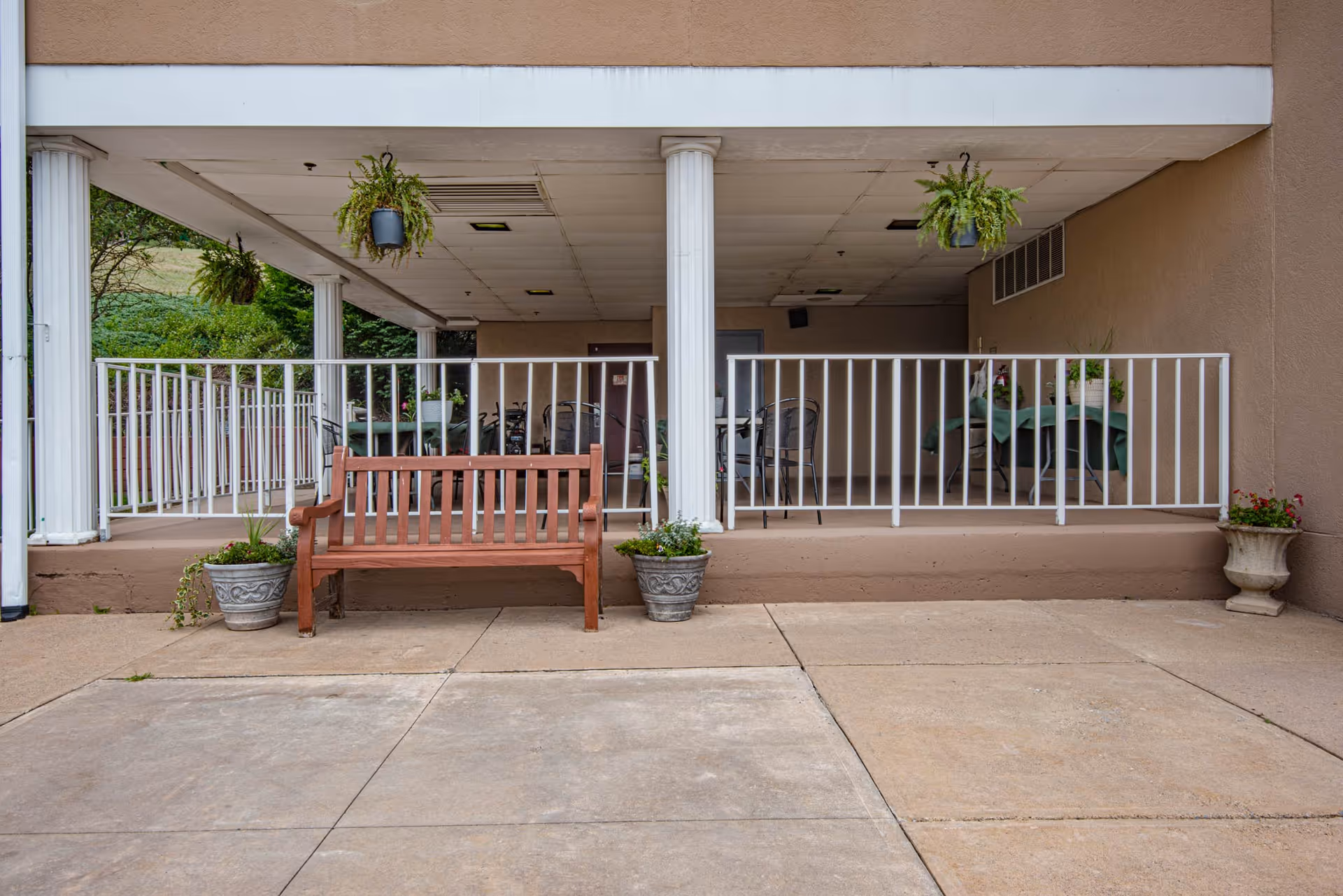 Covered outdoor patio area with white railings and columns, a wooden bench flanked by two potted plants, hanging ferns, and tables with chairs inside the covered space.