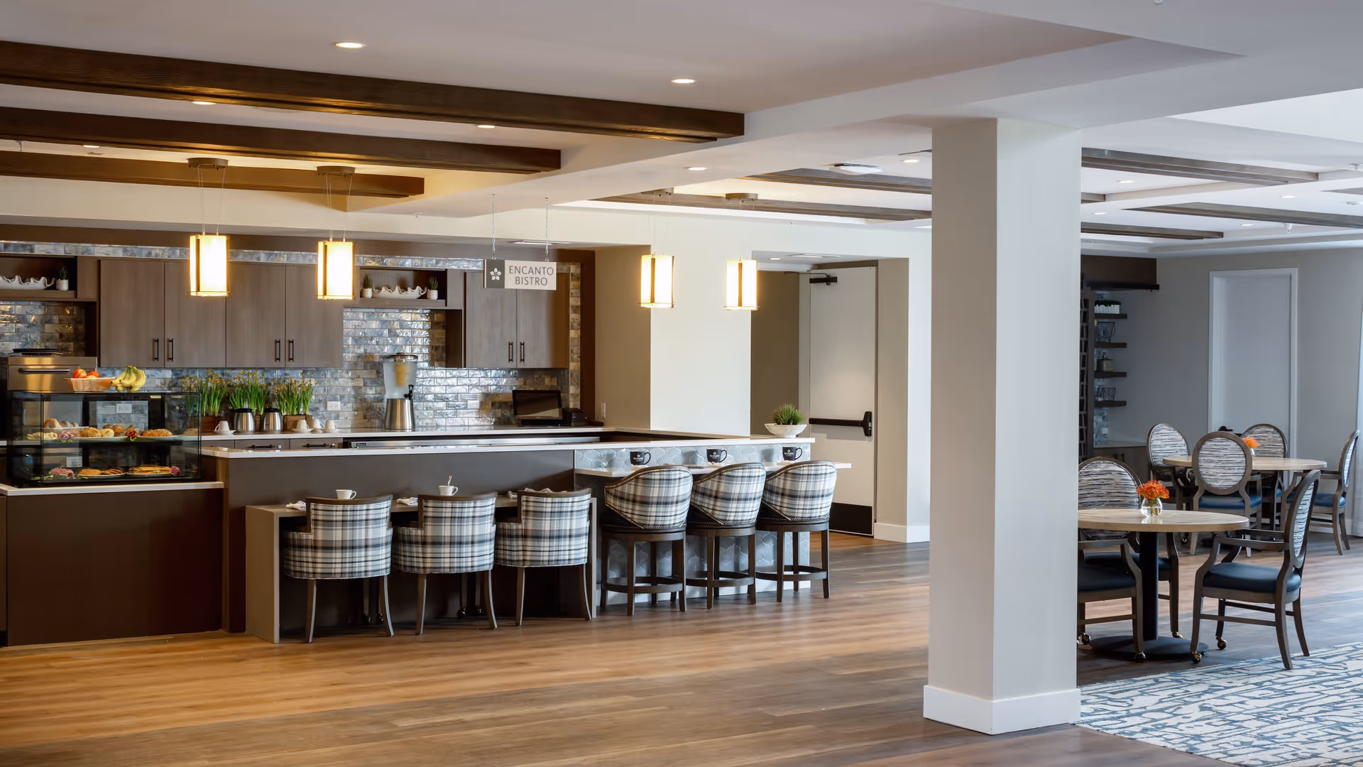 Interior view of a senior living facility dining area featuring a modern bistro counter with plaid cushioned chairs, pendant lighting, and a display case with pastries and fruit. There are also round tables with chairs and small flower arrangements, wooden ceiling beams, and a wood floor.