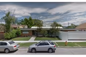 Street view of a single-story residential building with a flat roof, surrounded by trees and greenery. Two cars are parked on the street in front of the building, and a fire hydrant is visible on the sidewalk.