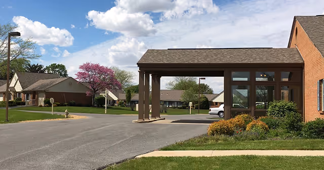Exterior view of a senior living facility with a covered entrance supported by columns, a driveway, landscaped bushes, and a background of residential-style buildings and trees under a partly cloudy sky.