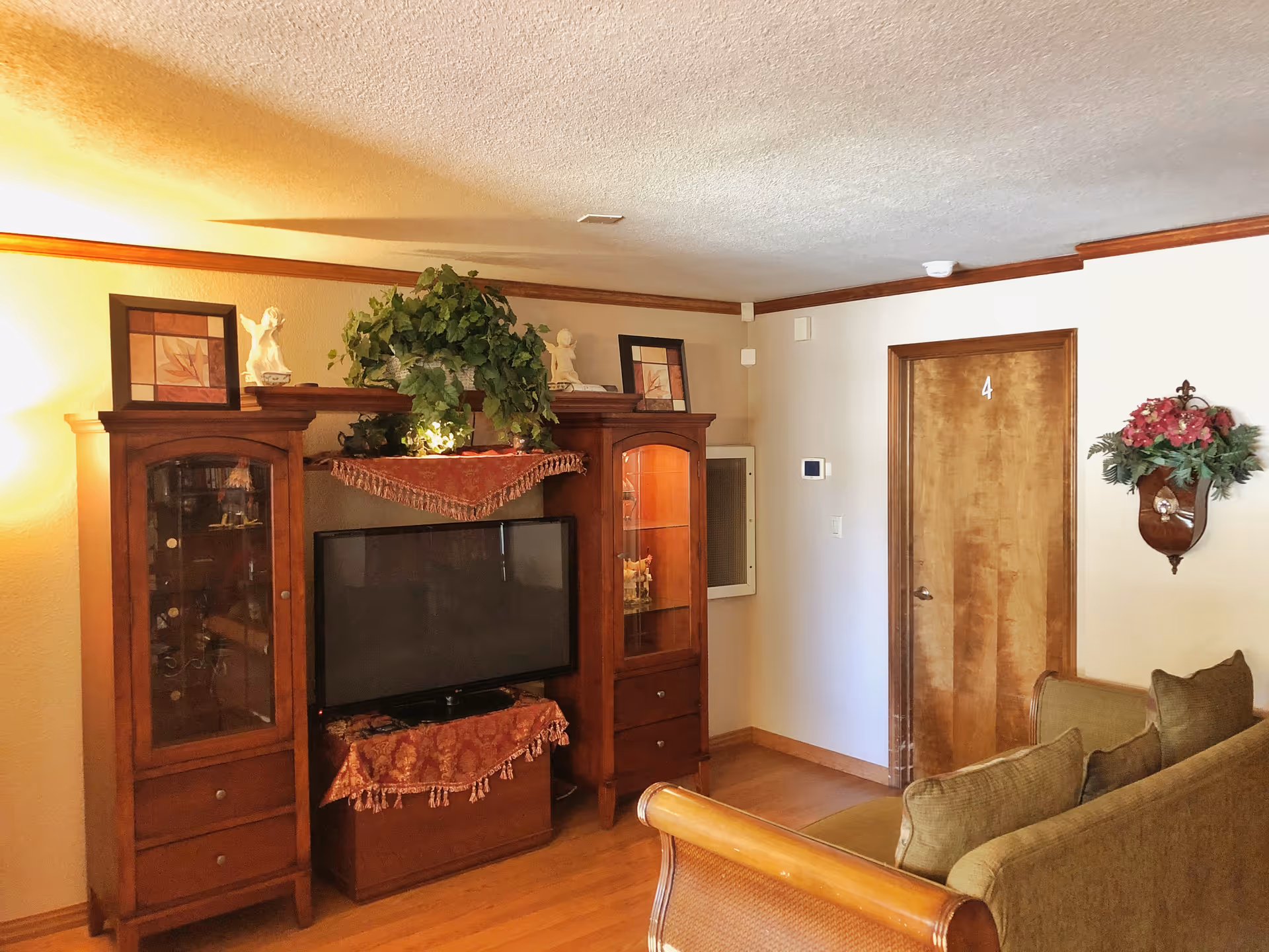 A cozy living room with wooden furniture including a TV cabinet with glass doors and drawers, a flat-screen TV, and a green couch with cushions. The walls are light-colored with wooden trim, and there is a wooden door labeled with the number 4. Decorative items such as framed pictures, small statues, a potted plant, and a floral wall hanging are visible.