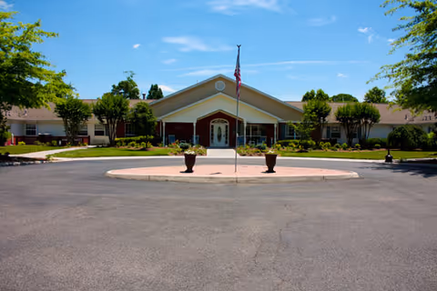 Front entrance of a single-story senior living facility with a circular driveway, American flag, and landscaped grounds under a clear blue sky.